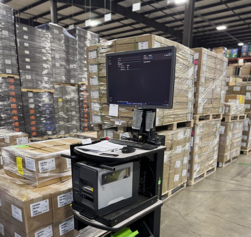 mobile-label-printing-workstation-inontime Mobile label printing workstation in a warehouse, positioned in front of stacked pallets with barcode-labeled boxes.