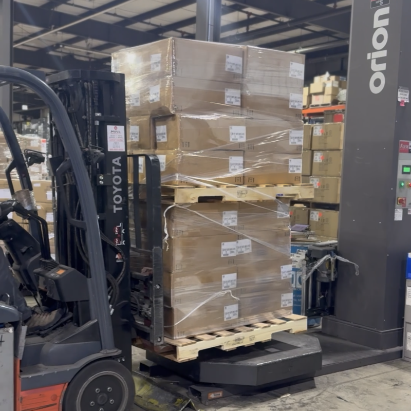 IMG_4778 Forklift moving a shrink-wrapped pallet of boxed goods onto an automated stretch wrapper in a warehouse.